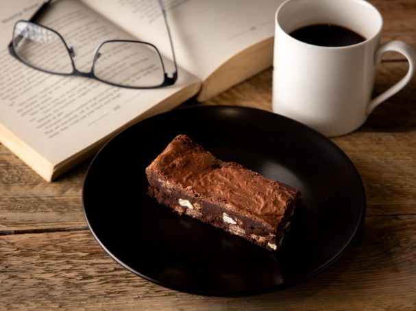 A chocolate brownie on a black plate beside a cup of coffee and an open book.