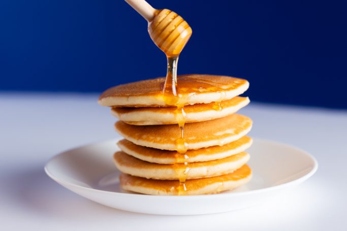 A stack of golden-brown pancakes sits on a white plate, Thick, amber-colored honey is being drizzled from above.