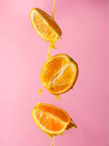 Sliced oranges hanging against a pink background with juice dripping down.