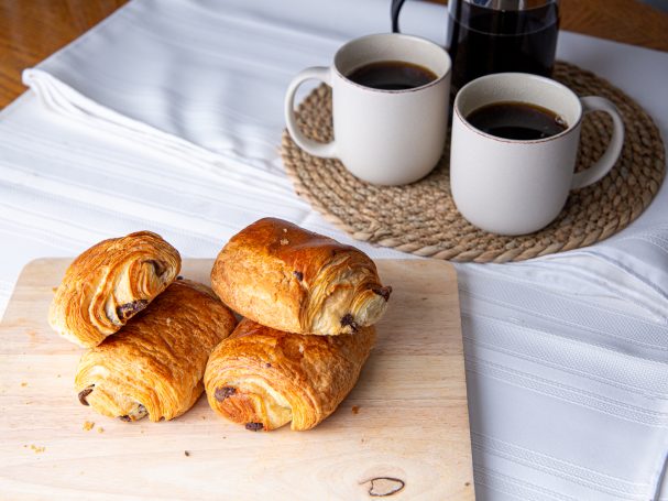 Four chocolate croissants on a wooden board with two cups of coffee nearby.