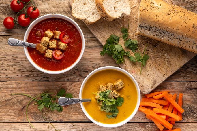 Two bowls of soup with croutons, surrounded by bread, tomatoes, carrots, and herbs.