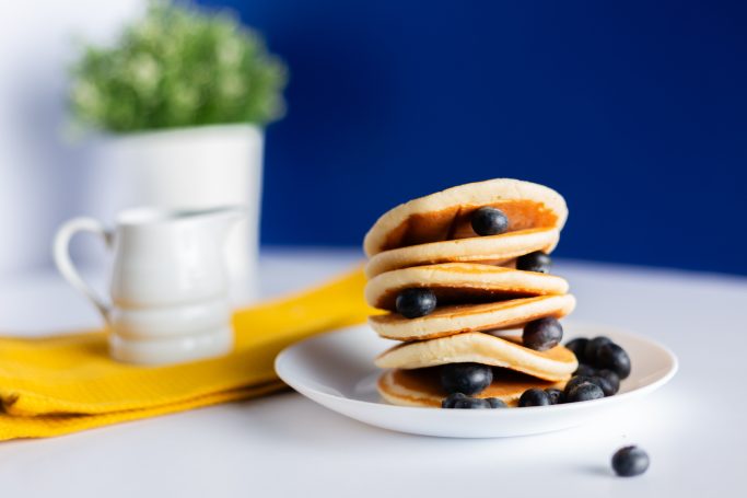 
A stack of fluffy, golden-brown pancakes is neatly arranged on a white plate, topped with a handful of fresh, plump blueberries. 