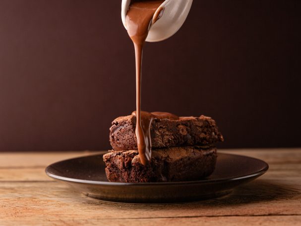 Rich chocolate sauce drizzling over two stacked brownies on a dark plate.